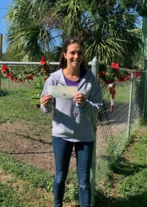 Michal Anne holding the check from SPCA of Manatee County, standing outside the Satchel's gate.