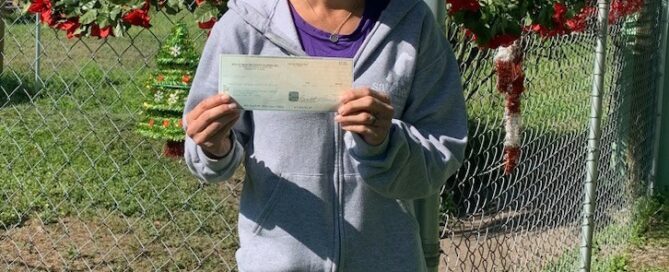 Michal Anne holding the check from SPCA of Manatee County, standing outside the Satchel's gate.