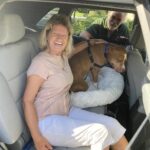 Cowboy sitting in the back of the vehicle with his new mom ready to head to his forever home. Dad staning beside.