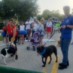 Trouble standing waiting for the parade to begin. Handler, Quentin, by his side. Volunteer and dog, Jorah, close by. Surrounded by many people waiting.